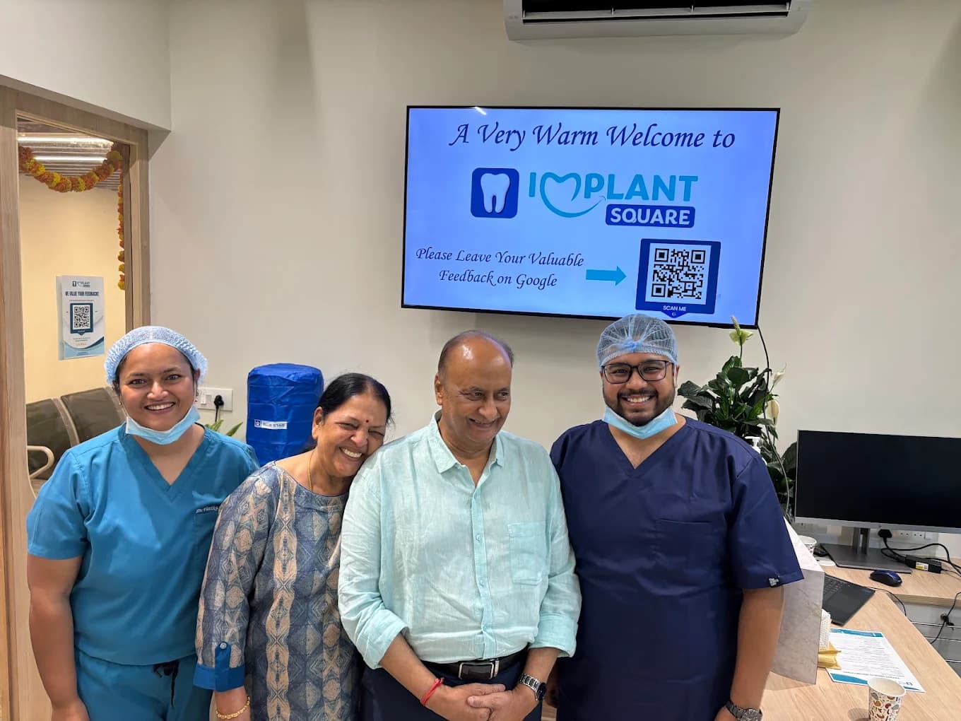 Implant Square Dental staff in scrubs smiling with two patients in the reception area, welcome screen and Google review QR code visible on the wall.